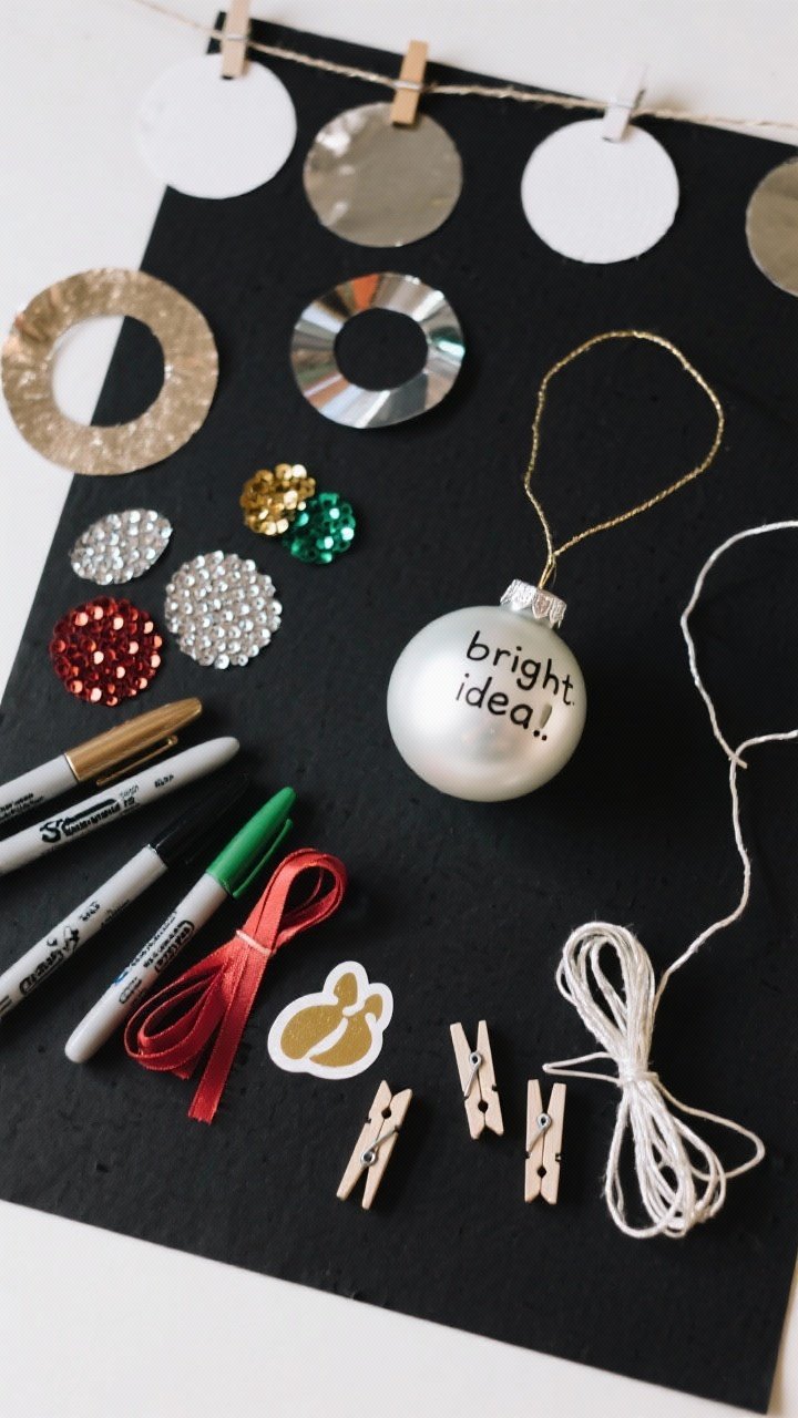 Overhead detail shot of ornament-making elements before being hung: deep black paper background sample, pre-cut ornament circles, metallic markers, sequins, stickers, ribbon lengths, and mini clothespins next to a thin garland string; one finished ornament labeled with a student’s name and a “bright idea” message; bright, even top lighting to highlight shine and color; tight composition focusing on textures and materials.